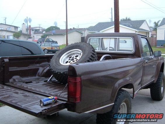 Brown pickup truck bed with a spare tire and a can of spray paint. Other vehicles and houses in background
