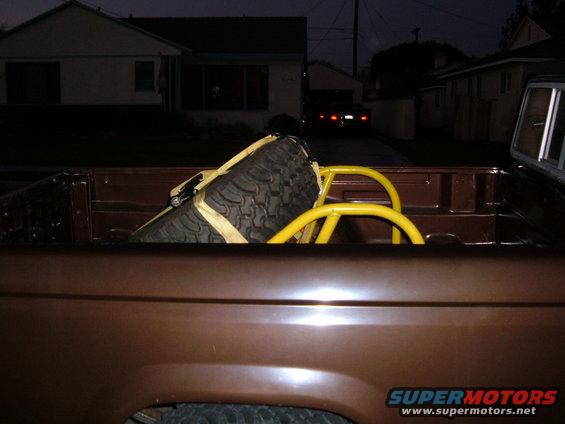Brown pickup truck bed with a spare tire secured by yellow straps and a yellow roll bar