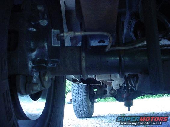 Underside view of a vehicle's rear axle and suspension components. Shows brake lines and part of a tire