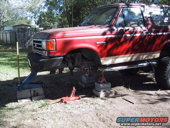 Red 1990 Ford Bronco with tan stripe, on jack stands, front 3/4 view