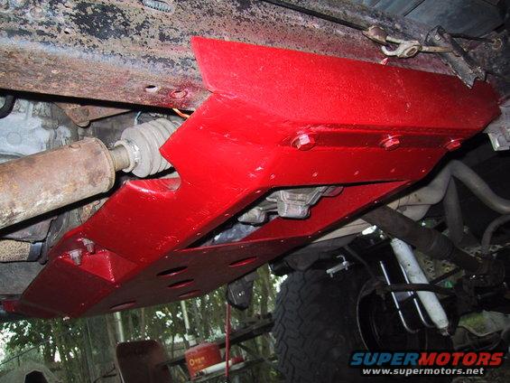 Underside view of a red skid plate bolted to a vehicle frame, showing drivetrain components and suspension