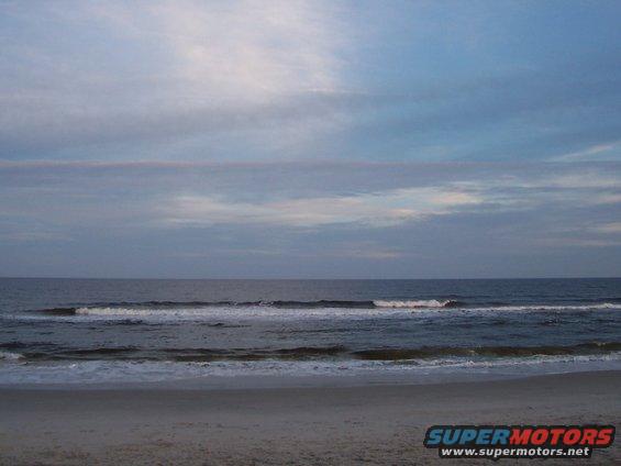 Beach with ocean waves under a cloudy sky, and a Supermotors.net watermark
