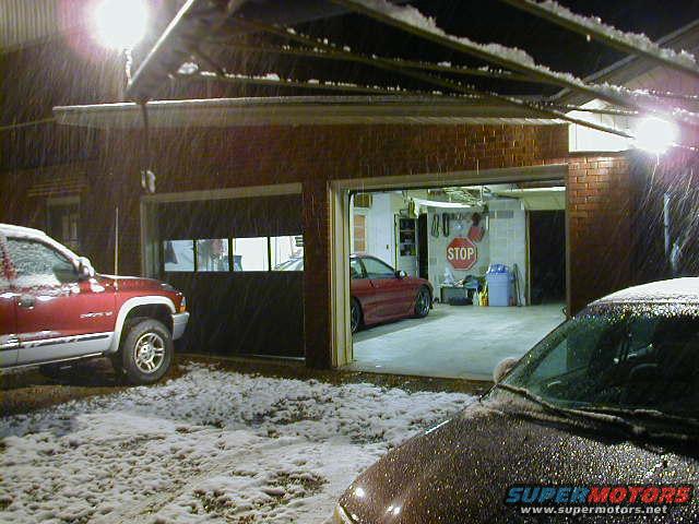 Red pickup truck and a red sports car parked near a garage with snow falling
