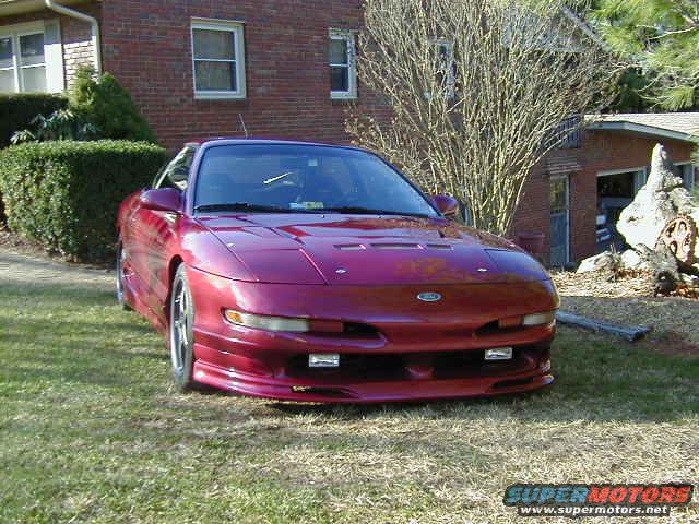 Red 1994 Ford Probe parked on grass in front of a brick house. The car has a sporty body kit and aftermarket wheels