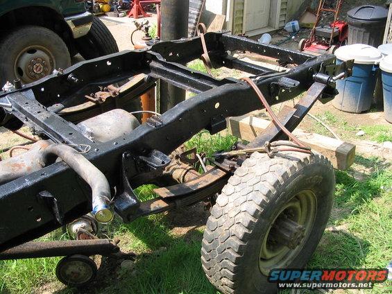 Truck frame with suspension components and a tire. Another vehicle is partially visible in the background