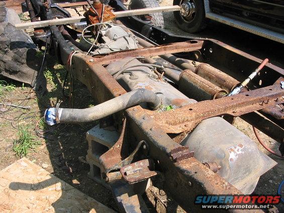 Heavily rusted truck frame with fuel tank and exhaust pipes visible; a wooden plank rests on top