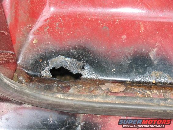 Close-up of rust hole in red vehicle body panel near a rubber seal