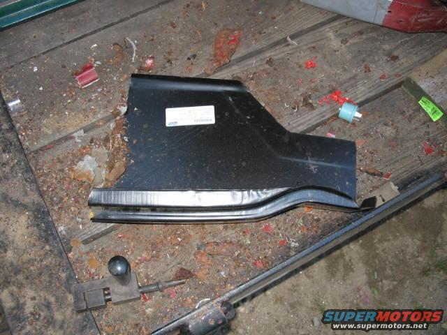 Black metal car rocker panel with a white sticker, lying on a wooden surface with debris