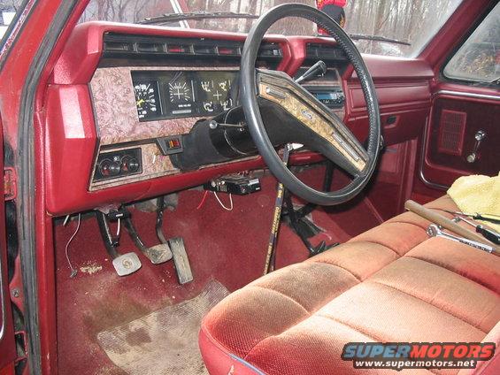Red truck interior with wood-grain dashboard, analog gauges, and worn fabric seats. Tools are on the passenger seat