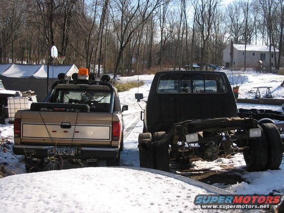 Rear view of a tan pickup truck next to a black truck chassis in a snowy yard