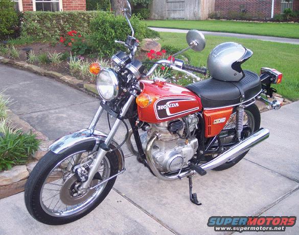 Red 1974 Honda CB360T motorcycle parked on a sidewalk, with a silver helmet on the seat