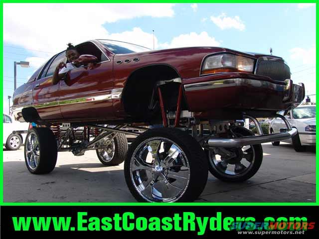 Maroon sedan with extreme hydraulic lift, chrome wheels, and a person in the driver's seat. Another white car is visible in the background