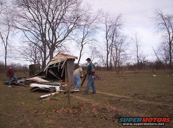 People are cleaning up debris from a collapsed wooden structure in a field with bare trees