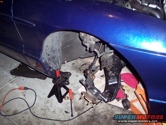Blue car's front suspension disassembled on jack stands, with "SUPERMOTORS.NET" visible