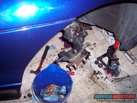 Disassembled car suspension components on the floor next to a blue car fender and a bucket