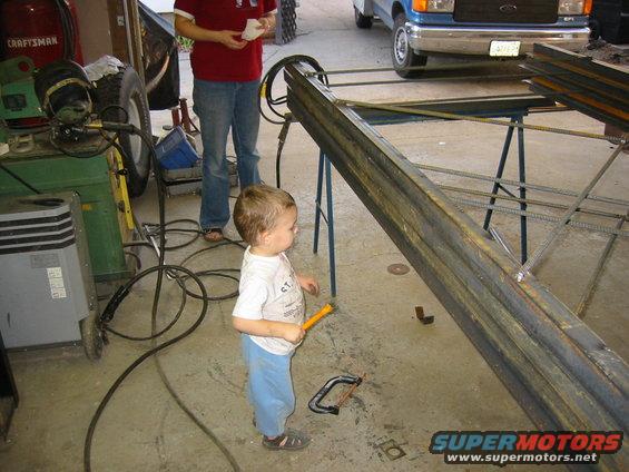 Kid watching metal beams being welded in a garage, blue Ford van in background