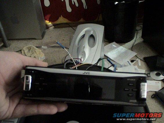 JVC car stereo with wires attached, held in hand. Background shows speakers and papers