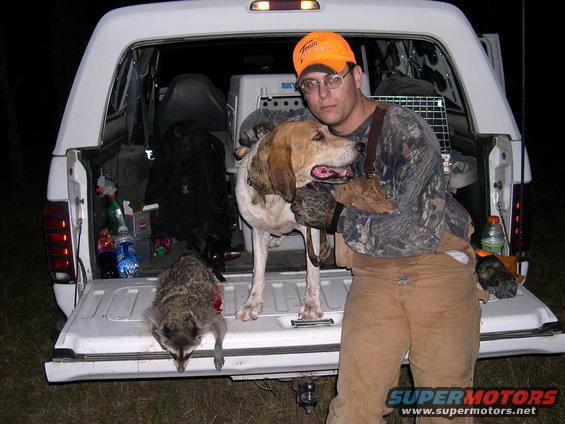Man in camouflage holding a hound dog, with a raccoon in the open tailgate of a white pickup truck