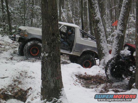White off-road truck with large tires navigating a snowy forest, driver visible