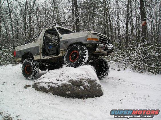 Silver modified truck with large tires on a snowy rock, door removed, roll cage visible
