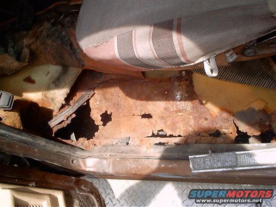 Passenger side floor pan of a vehicle with severe rust damage and holes, showing the seat base above