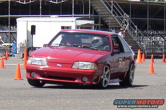 Red 1987 Ford Mustang hatchback with racing cones on track. Driver wears helmet