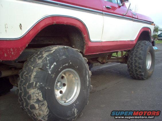 Red and white Ford Bronco with large off-road tires, lifted suspension, and chrome trim. Visible rust spots on the white panel