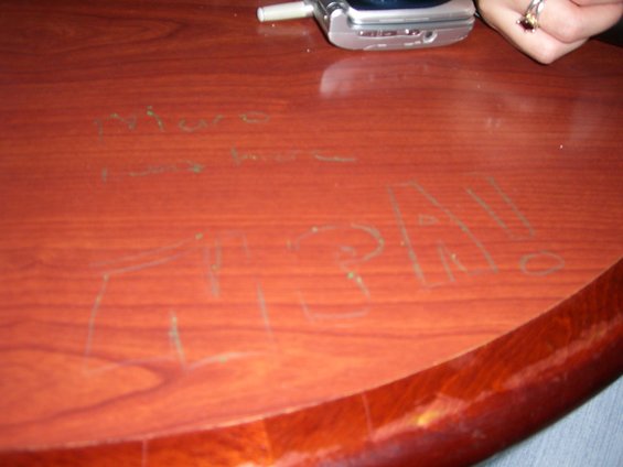 Graffiti scratched into a wooden table surface, with a cell phone and a hand visible