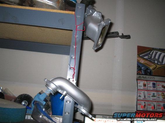 Silver metal engine intake manifold hanging from a gray metal strip with red wire