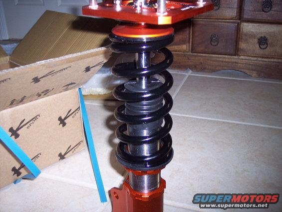 Adjustable coilover suspension part with black spring and orange mounting bracket. Cardboard box with 'SPORT' logo visible