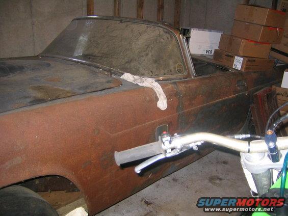 Rusty 1956 Ford Thunderbird side view, showing a cloth on the roof and a motorcycle handlebar in the foreground