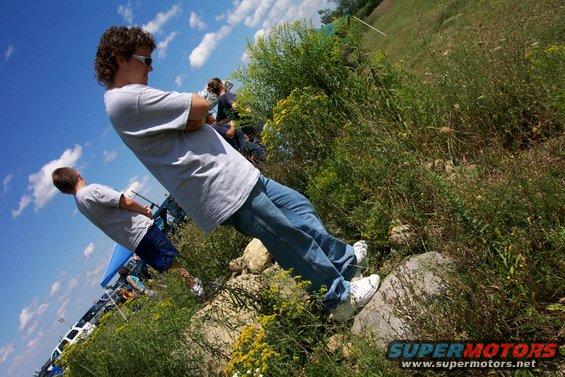 Two guys standing in a field with a blue tent and some vehicles in the background