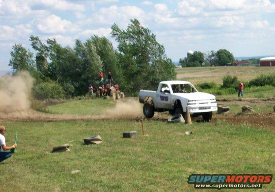White pickup truck with large tires navigating an off-road course, kicking up dust