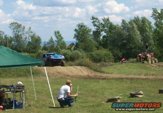 Blue off-road truck climbing a dirt mound, people watching from a grassy area