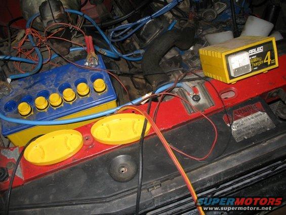Engine bay with a blue and yellow battery, yellow battery charger, and red and black wires. A "WARNING" sticker is visible