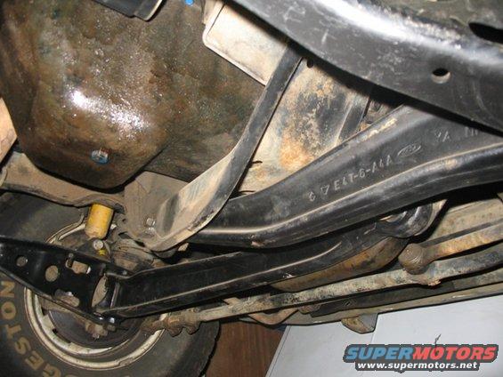 Underside of a vehicle showing suspension components and part of a tire. A Ford logo and part number are visible on a suspension arm