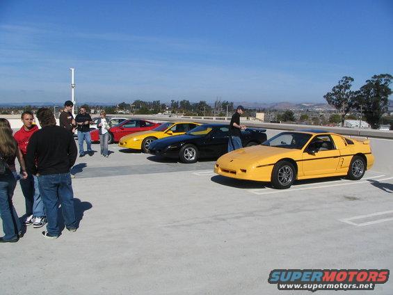 Yellow 1986 Pontiac Fiero parked in front of other Fieros and people