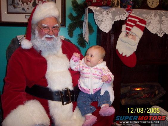Santa Claus in his suit holding a baby girl wearing a pink and white sweater and blue jeans