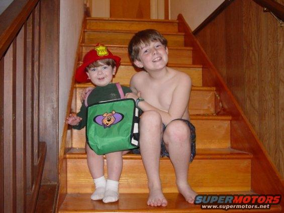 Two young boys sit on wooden stairs; one wears a firefighter hat and carries a green bag with a cartoon bear