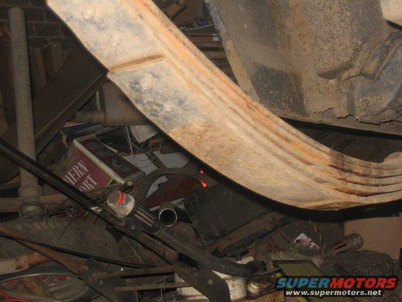 Underside of a vehicle showing a rusty leaf spring and exhaust pipe, with various parts and a sign in the background