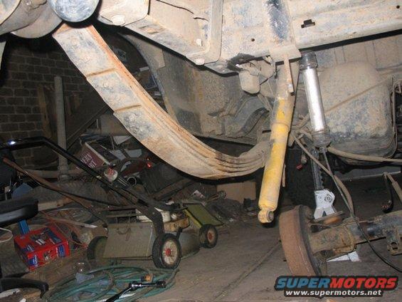 Leaf springs and shock absorber on a vehicle's rear axle, with a lawnmower and other items in the background