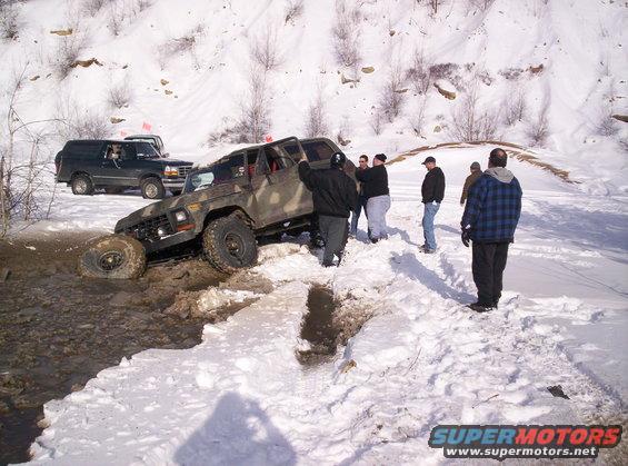 Dark green 1980s Ford Bronco parked in snow next to a camouflage off-road vehicle stuck in mud