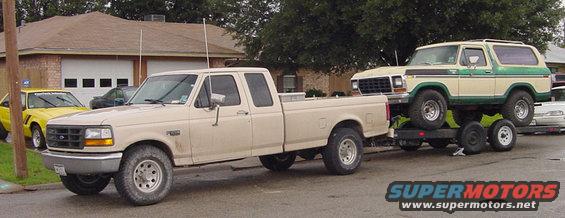 1978 Ford Bronco on a trailer, towed by a beige Ford F-150 pickup truck. Yellow car in background