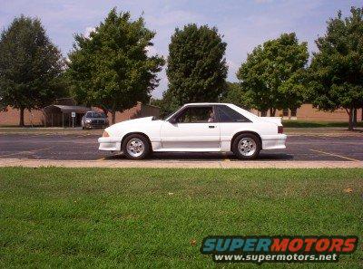 White coupe parked on asphalt with grass in foreground and trees in background