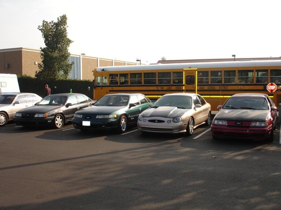 Four sedans parked in a lot, with a school bus behind them. The closest car is a red Ford Taurus