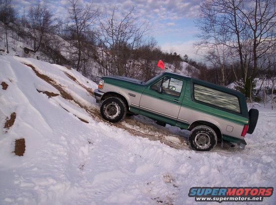 1996 Ford Bronco driving up a snowy, rutted hill with trees in the background