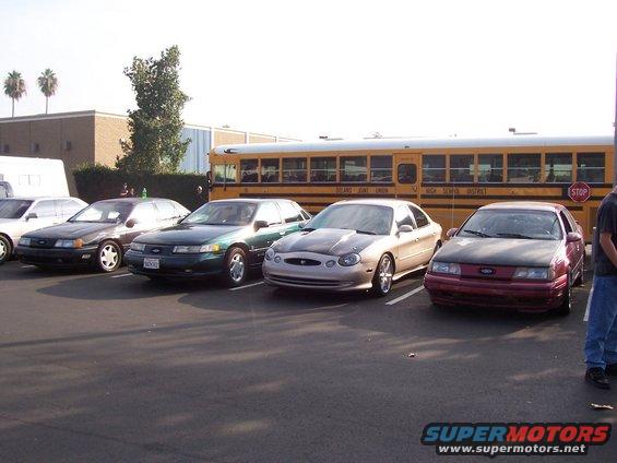 Four sedans parked in a lot, one is a maroon 1996 Ford Taurus with a dark hood, another is a silver 1996 Ford Taurus with a dark hood, and a green sedan is visible