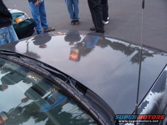 Dark colored car hood with reflections of people and a traffic light; windshield wipers visible