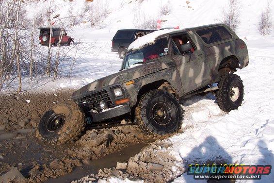 Camouflaged Ford Bronco stuck in mud and snow, front-left wheel deep in mud