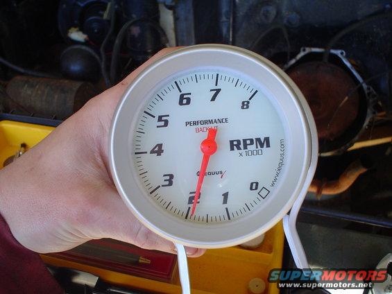 Hand holding a tachometer gauge with red needle pointing to 2